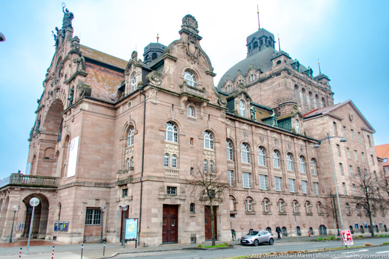 Oblique street view of the sandstone Opernhaus with tall arched windows, gables, and a high roof mass.
