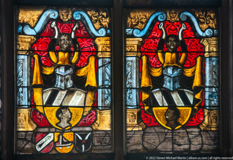 Two adjacent heraldic stained-glass panels with shields, dark supporting figures, red canopies, and gold ornament.
