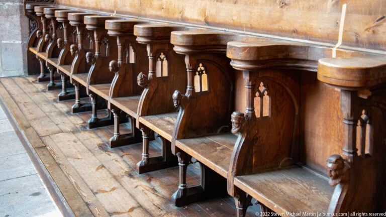 Row of dark wooden choir stalls with carved dividers and small sculptural details along a stone floor.