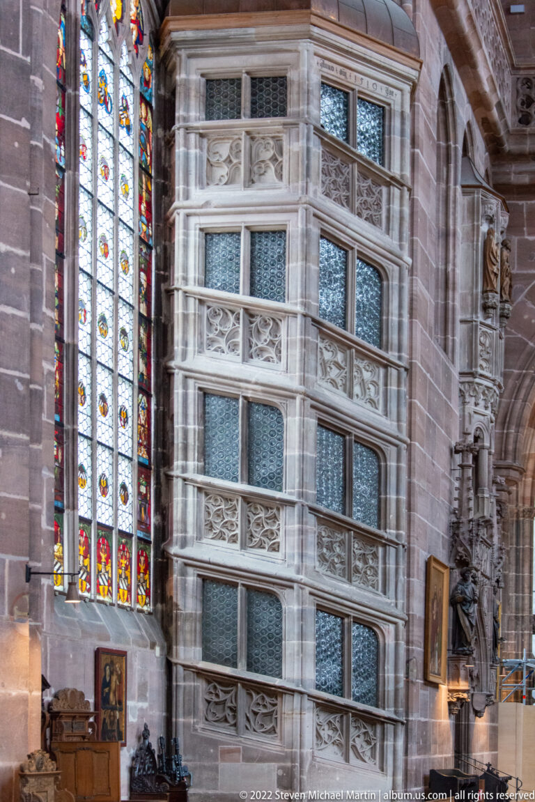 Tall pale stone enclosure with framed openings and carved tracery beside a stained-glass window inside St. Lorenz.