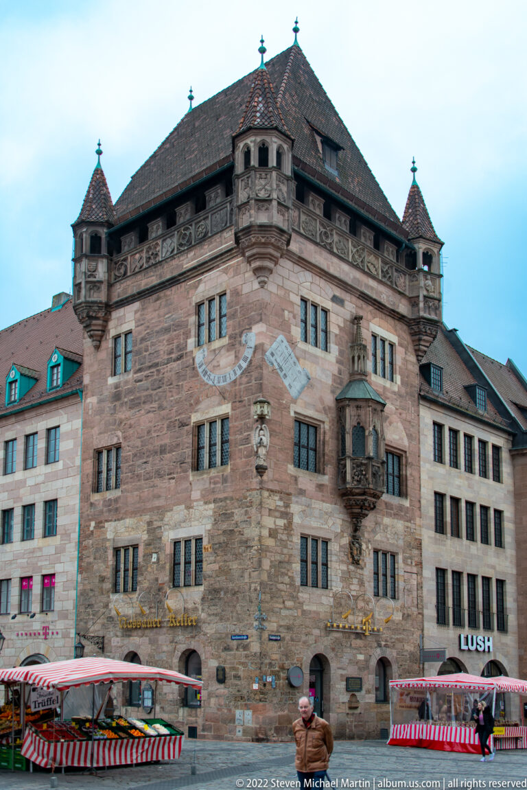 Large sandstone civic building with painted sundial, ornate corner oriel, hipped roof, and market stalls below.