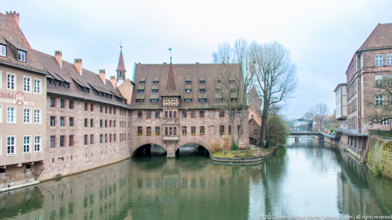 Heilig-Geist-Spital spanning the Pegnitz on masonry arches, with pitched roofs and reflections in the river.