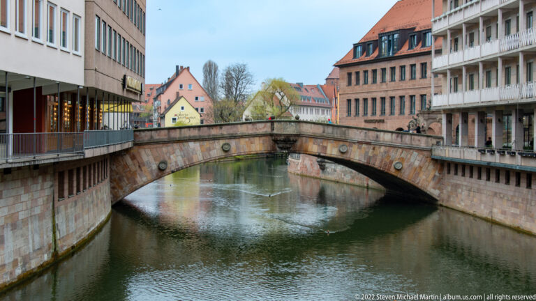 Single-arch stone bridge over the Pegnitz between closely spaced buildings and riverside walkways.
