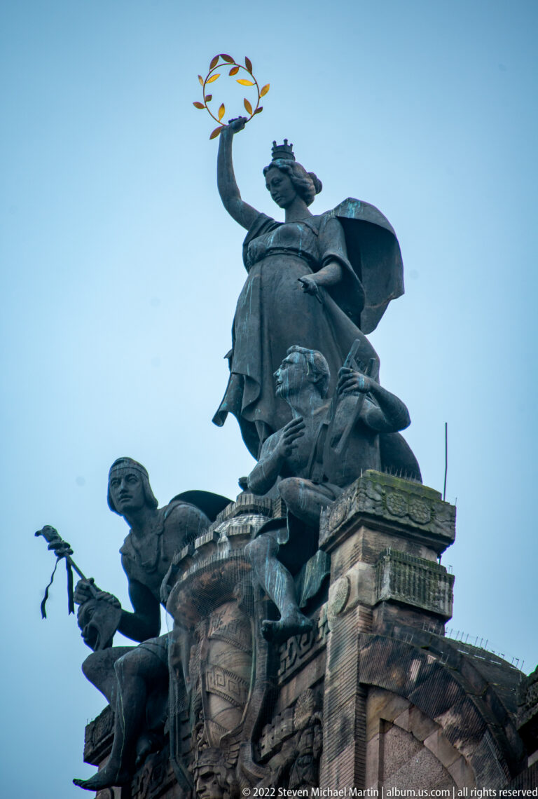 Dark rooftop sculptural group with a seated figure raising a gilded wreath above smaller figures.