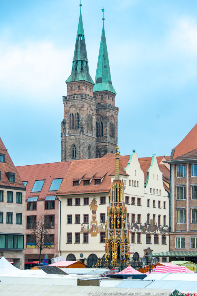 Schöner Brunnen rising above market stalls with stepped-gable buildings and St. Sebald’s twin towers behind.
