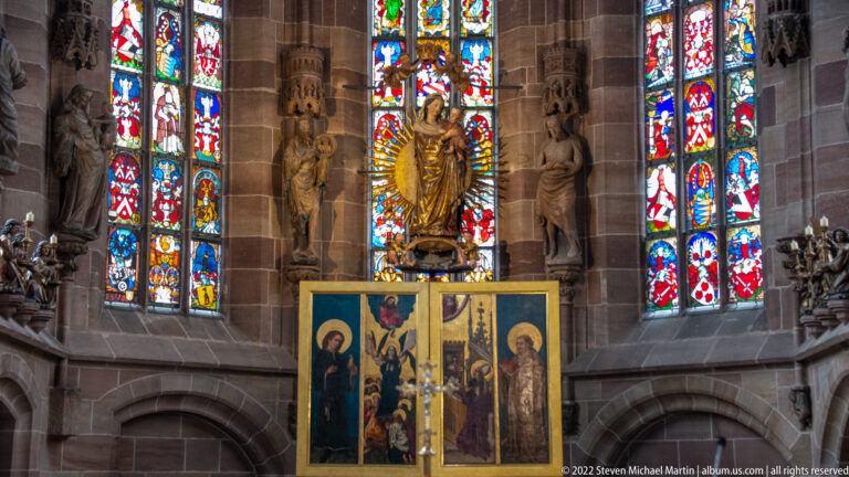 Crowned Madonna and Child with radiant halo above a painted triptych, framed by stained glass and stone figures.