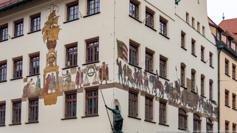 White historic building with painted procession mural, heraldic motifs, red-trimmed windows, and a bronze statue below.