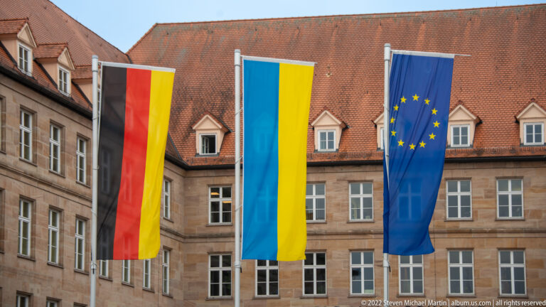 German, Ukrainian, and European Union flags hanging vertically before a historic stone building.