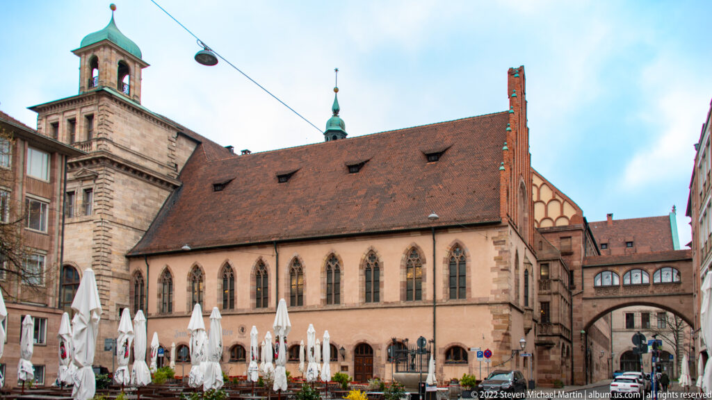 Long sandstone Gothic hall with pointed windows, steep tiled roof, café umbrellas, and an arched passage at right.
