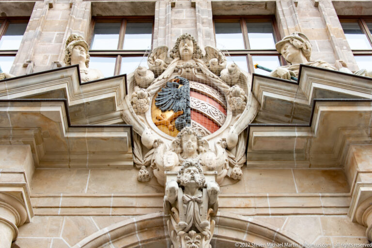 Ornate carved coat of arms with divided shield, putti, masks, and scrollwork above a stone portal.