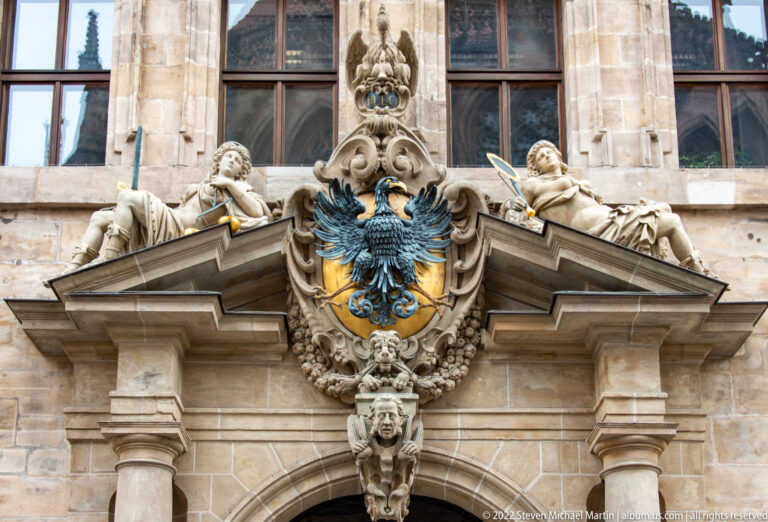 Black double-headed eagle on a gold shield surrounded by carved figures and scrollwork above a portal.