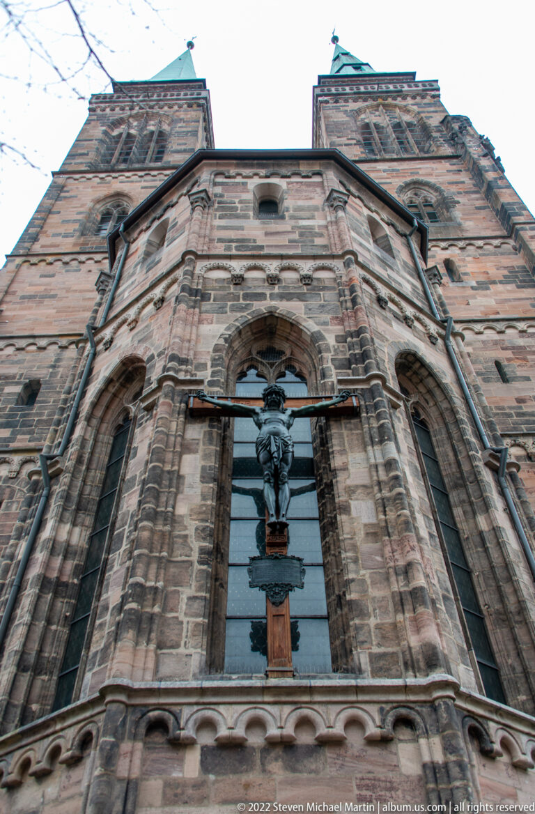 Dark crucifix mounted against a tall glazed opening in the sandstone exterior wall of St. Sebald Church.
