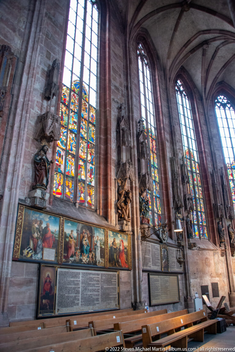 Painted epitaphs and memorial panels beneath tall stained-glass windows along a side aisle of St. Sebald.