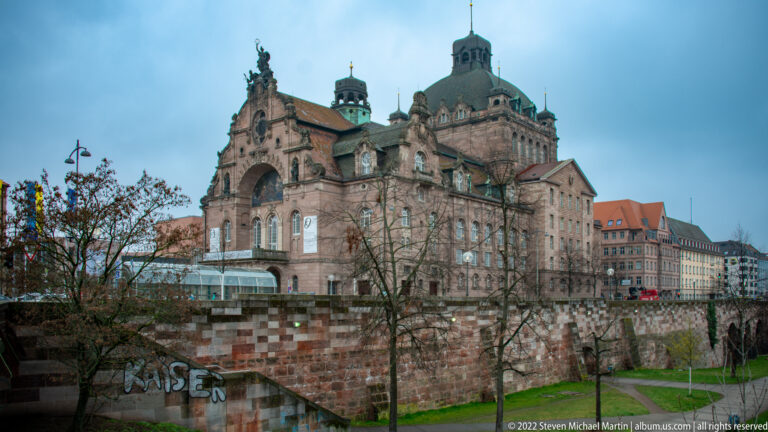 Nuremberg State Theatre rising beyond a brick retaining wall, trees, and a narrow landscaped path.