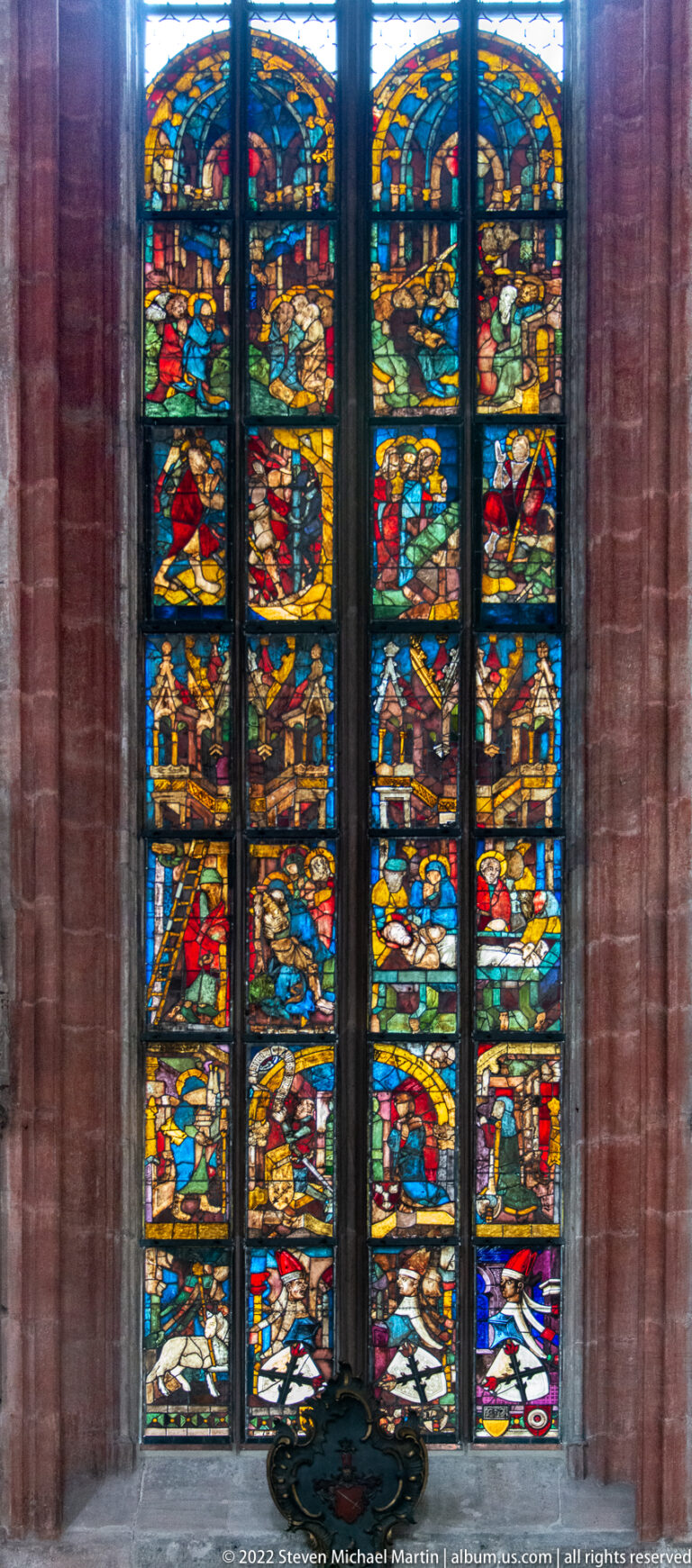 Close view of a tall stained-glass window with stacked biblical scenes in vivid red, blue, green, and gold glass.