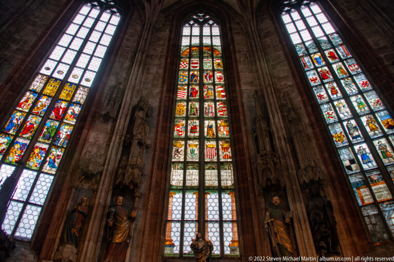 Three tall stained-glass windows between massive sandstone piers, viewed upward inside St. Sebald.