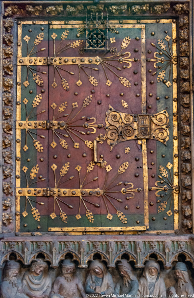 Green and purple decorative panel with gilded fleur-de-lis, metal bands, heraldic motifs, and carved detail below.