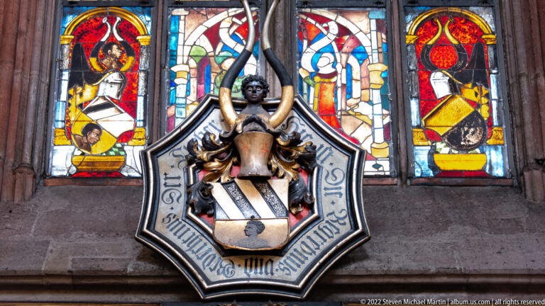 Large heraldic shield with striped coat of arms, crested helmet, mantling, and stained glass behind.