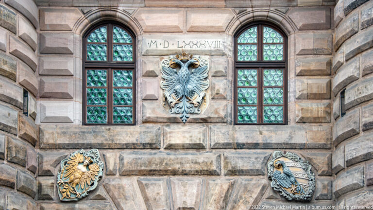 Double-headed eagle relief and two smaller heraldic shields between leaded windows on a rusticated stone wall.