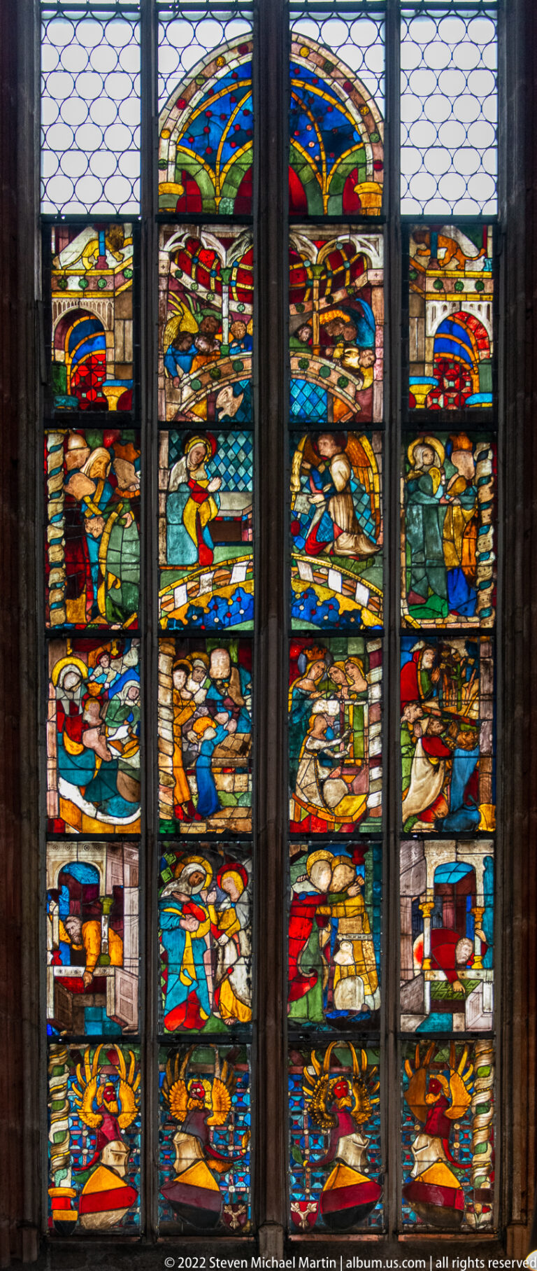 Multi-lancet stained-glass window filled with stacked colorful narrative scenes divided by dark mullions.