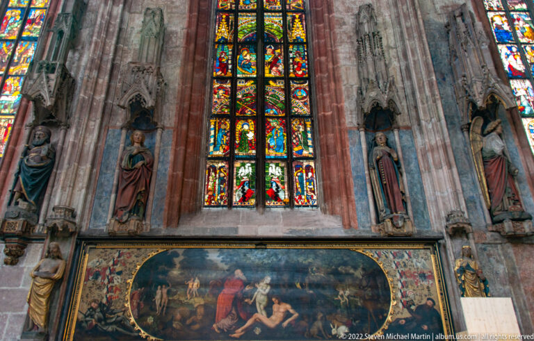 Tall stained-glass window above a broad narrative painting, flanked by carved stone figures in niches.