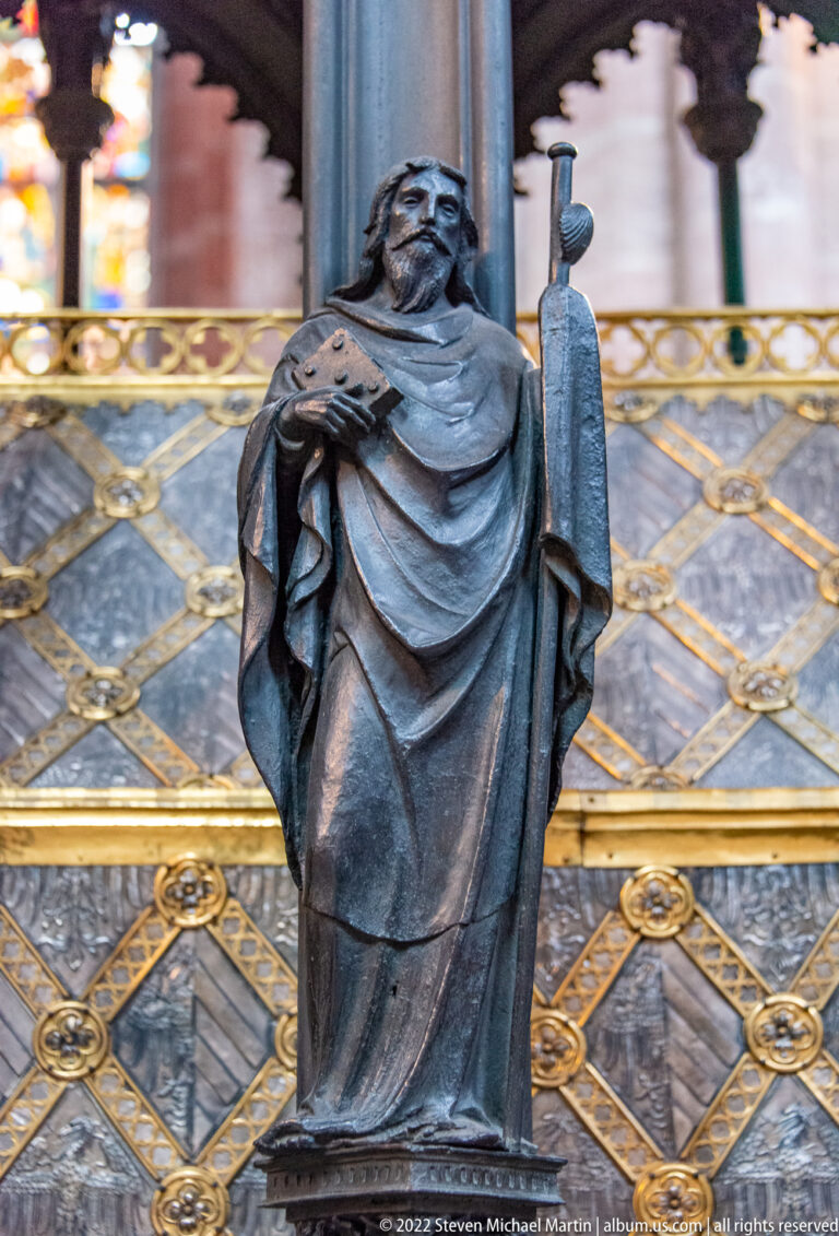 Small bronze robed figure holding a long attribute against the patterned metalwork of Saint Sebald’s tomb.