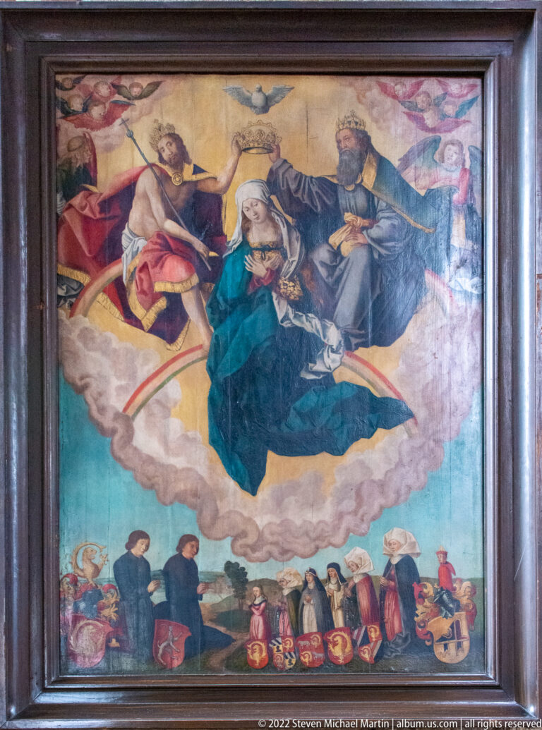 Framed religious painting with large sacred figures above kneeling donors and a row of heraldic shields below.