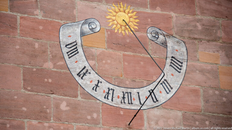 Horseshoe-shaped wall sundial with Roman numerals, diagonal metal gnomon, and golden sun motif above.