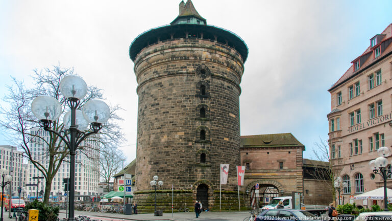 Round stone Frauentorturm with small openings and a broad conical roof beside a modern city street.