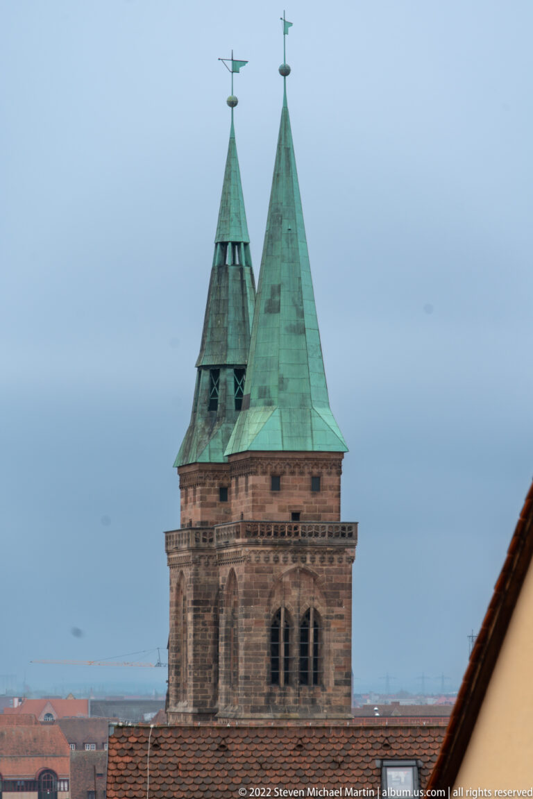 Square sandstone castle tower with paired green pyramidal rooflets, weather vanes, and narrow arched openings.