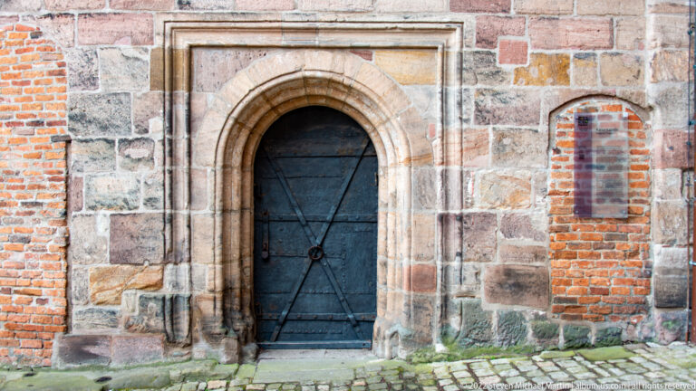 Round-arched stone doorway with dark iron-strapped wooden door set in mixed sandstone and brick castle masonry.