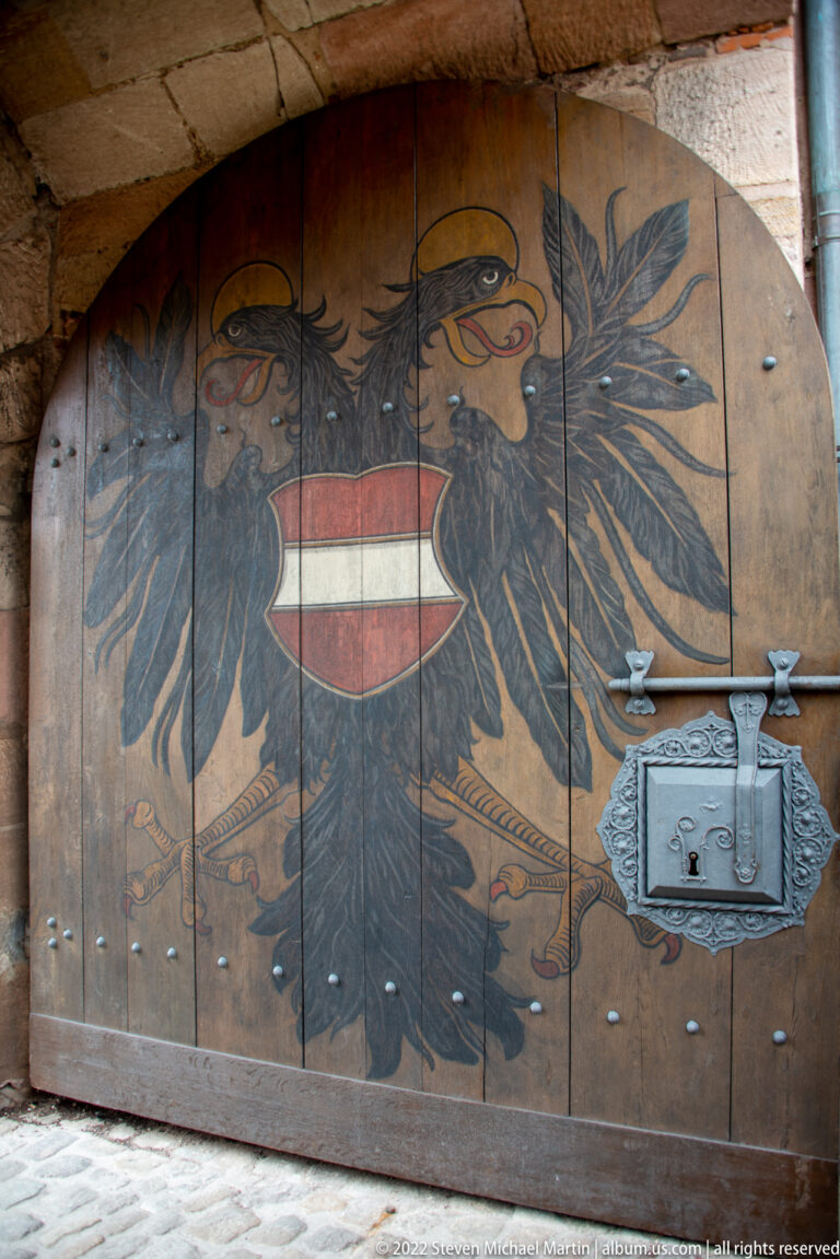 Black double-headed imperial eagle with red-and-white shield painted across a heavy wooden gate with iron lock hardware.
