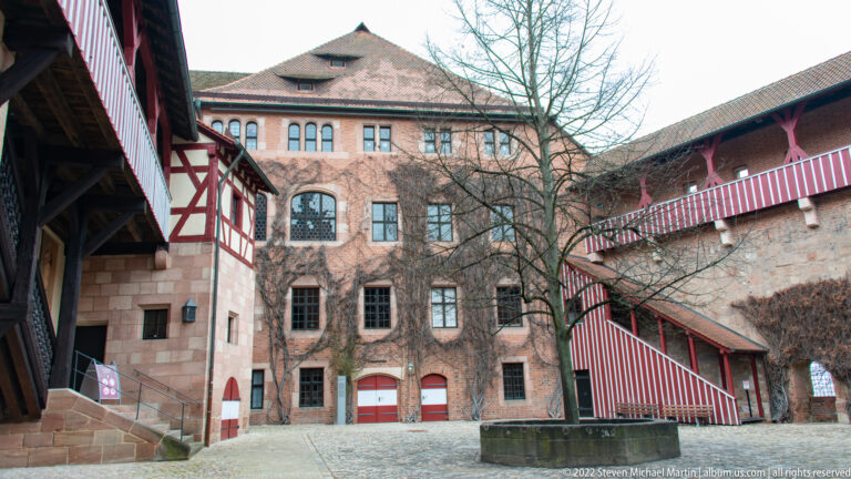 Imperial Castle courtyard with sandstone buildings, covered timber galleries, red stairways, and cobblestone paving.