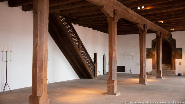 Interior castle hall with massive timber posts and beams, central wooden stair, white walls, and stone floor.