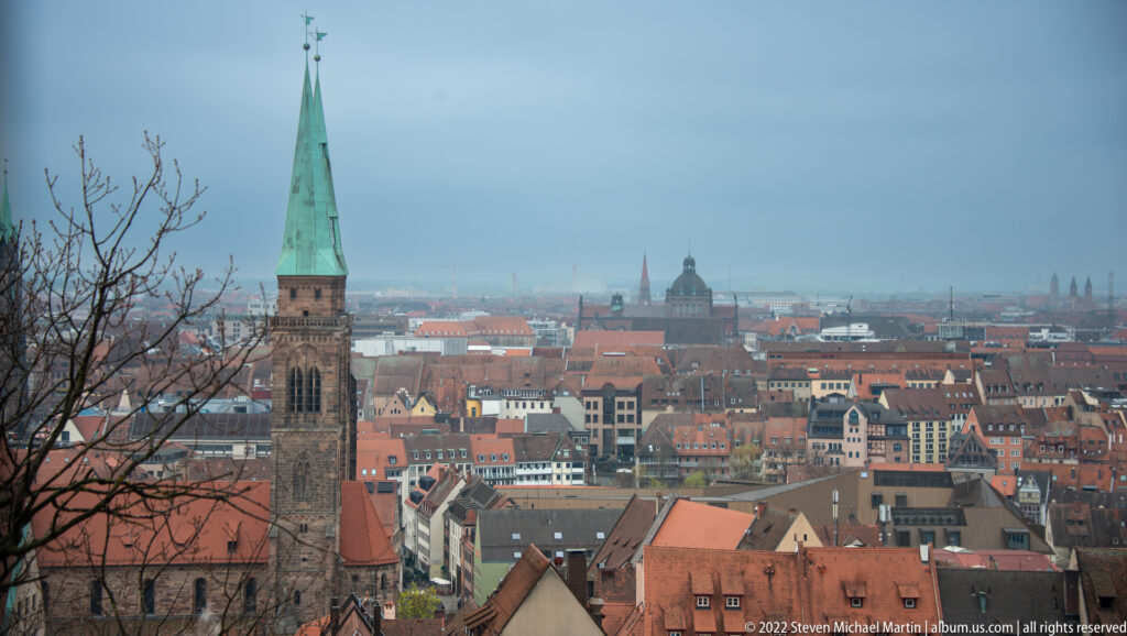 Elevated view over Nuremberg rooftops with a tall green-spired stone tower in the foreground and city skyline beyond.