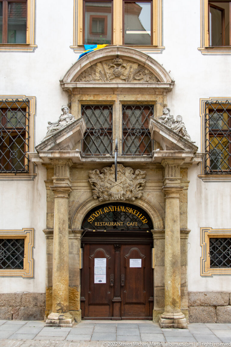 Elaborately carved stone doorway with columns and pediment between iron-grilled windows on a pale historic façade.
