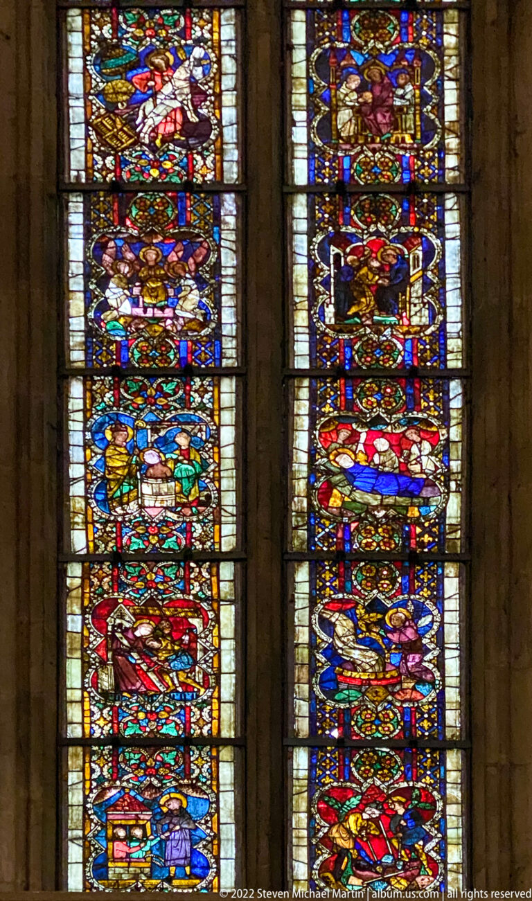 Two tall stained-glass windows with stacked figurative scenes in vivid red, blue, green, and gold glass.