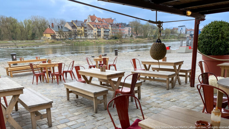 Outdoor riverside dining terrace with wooden tables, red chairs, and historic houses across the Danube.