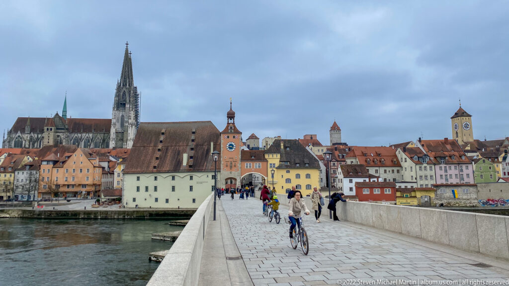 People and cyclists crossing Regensburg's Stone Bridge toward the old town, with St. Peter's Cathedral dominating the skyline.