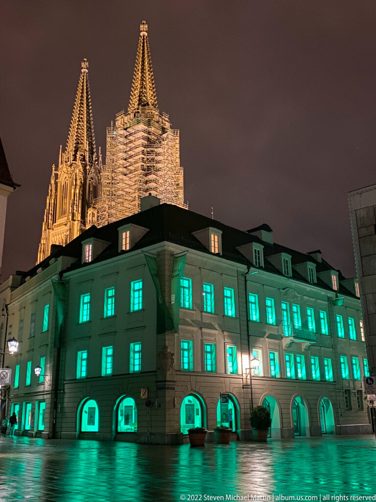 Green-lit historic building reflected in wet pavement, with illuminated Regensburg Cathedral towers rising behind at night.