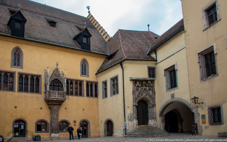 Historic courtyard with pointed-arch windows, a carved projecting bay, ornate doorway, arched passage, and visitors below.