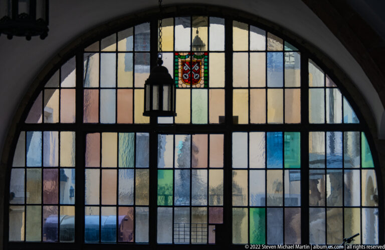 Large arched leaded-glass window with pastel panes, dark muntins, hanging lantern, and a small heraldic panel near the top.