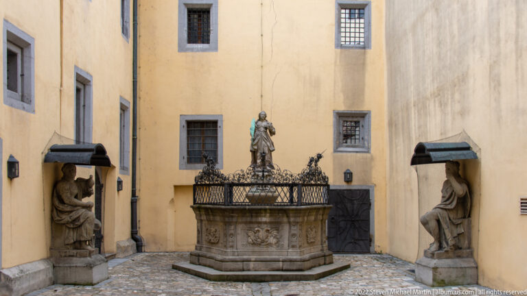 Stone courtyard fountain with a standing figure, ornate metal railing, and sculpted figures in wall niches on both sides.