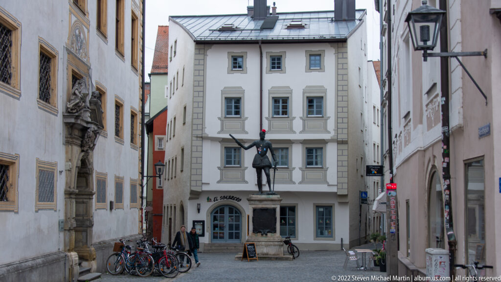 Bronze standing figure on a pedestal in a narrow Regensburg square, with bicycles and historic façades around it.