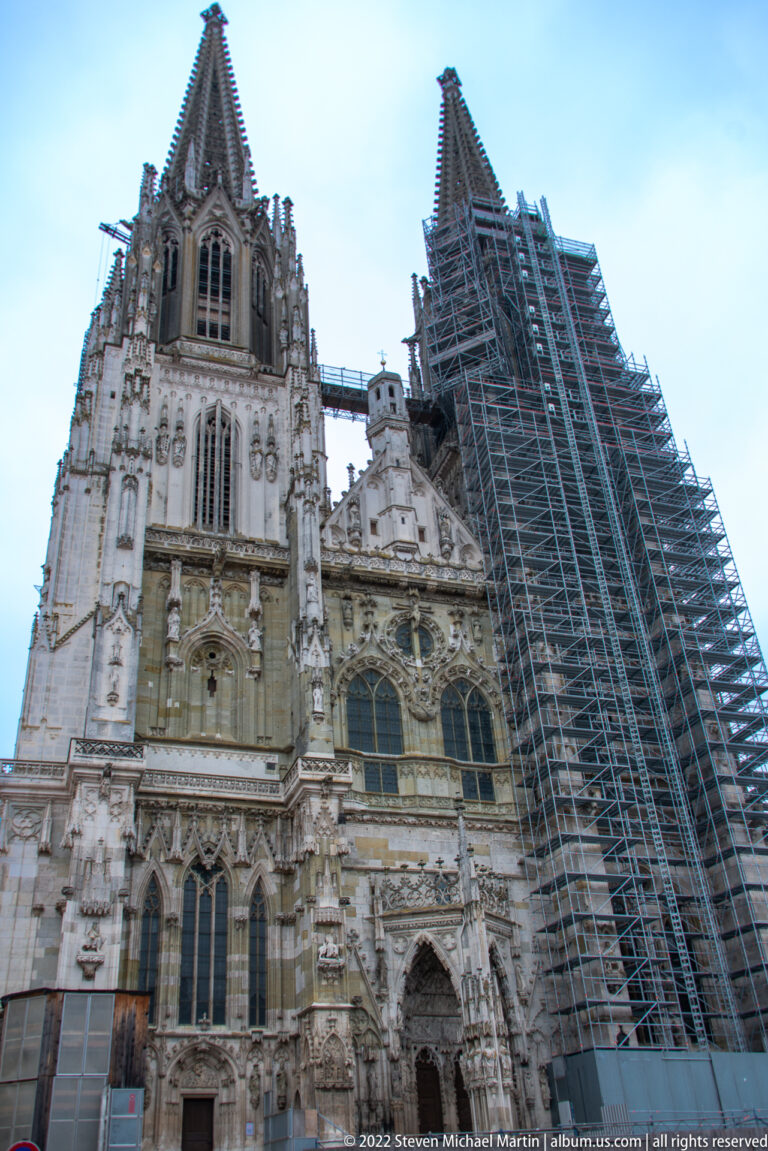 Ornate Gothic west front of Regensburg Cathedral with one tower heavily covered in scaffolding.