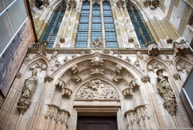 Upward view of an ornate Gothic cathedral portal with pointed arches, sculpted figures, relief carving, and tall windows above.