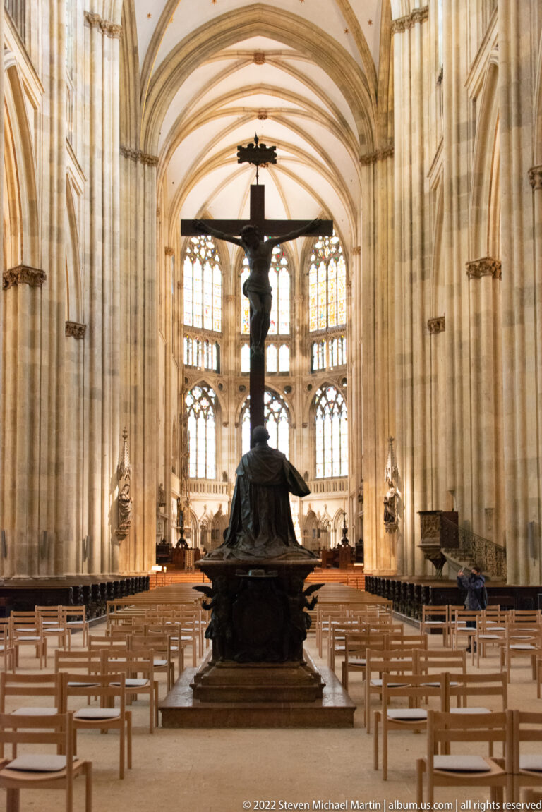 Central nave of Regensburg Cathedral with rows of chairs, a large crucifix monument, ribbed vaults, and bright choir windows.