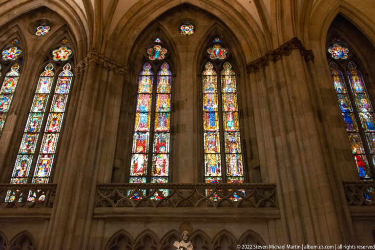 Three tall Gothic stained-glass windows with vivid multicolored panels and stone tracery inside Regensburg Cathedral.