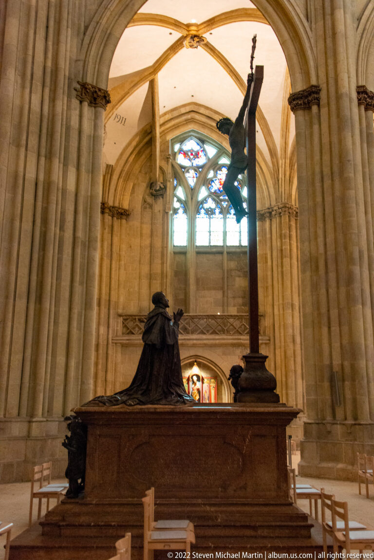 Side view of a large crucifix on a sculptural base before stone columns and stained glass in Regensburg Cathedral.