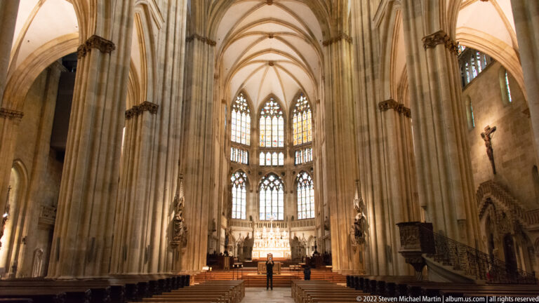 Long nave view inside Regensburg Cathedral with clustered stone piers, ribbed vaults, high windows, and the choir in the distance.